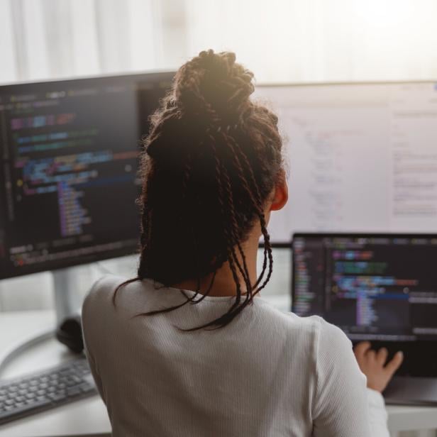 Rear of female web expert typing on computer looking at monitors while sitting indoors